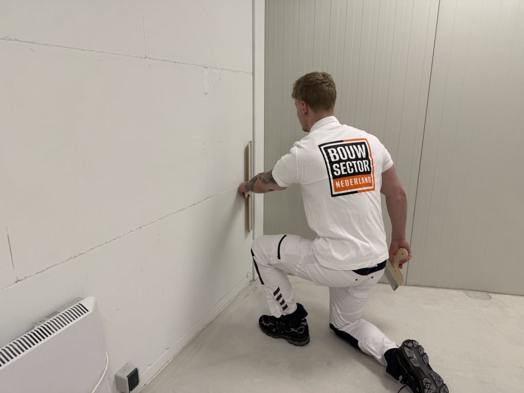 Knielende stukadoor in wit werktenue van Bouwsector Nederland trekt een rei langs de onderkant van een wand